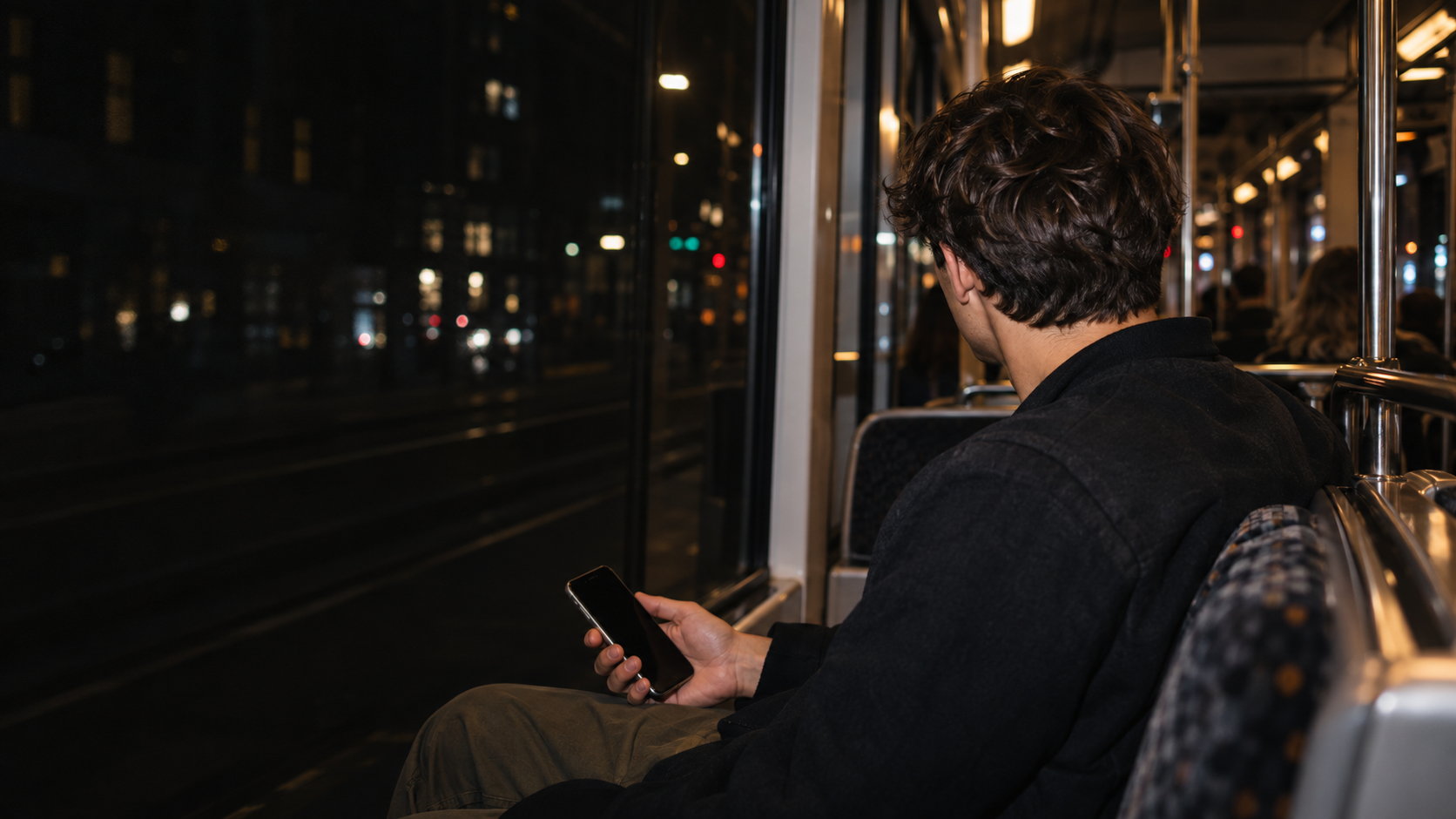 Jongvolwassene kijkt op van een gezakte telefoon in een tram bij nacht.